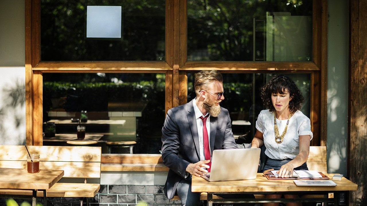 man in a suit and women with necklace outside a coffee shop with a women and laptop