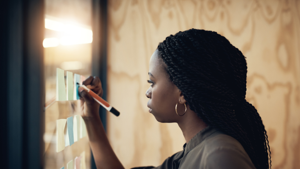 Female in business attire writing on different coloured post-it notes stuck to the glass wall of an office