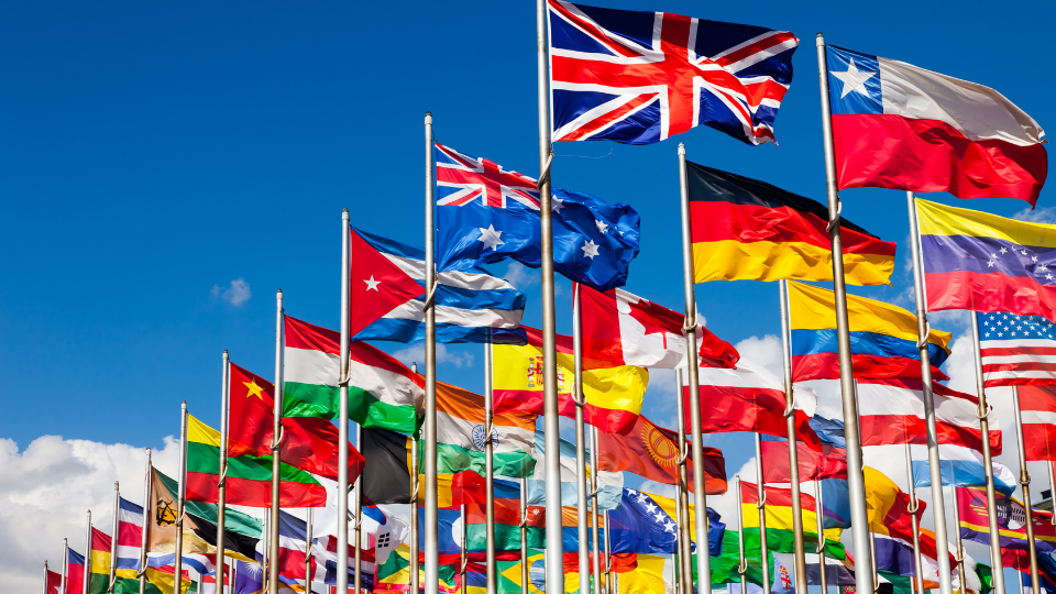 Lots of flags of countries of the world on flagpoles blowing in the wind against a blue sky