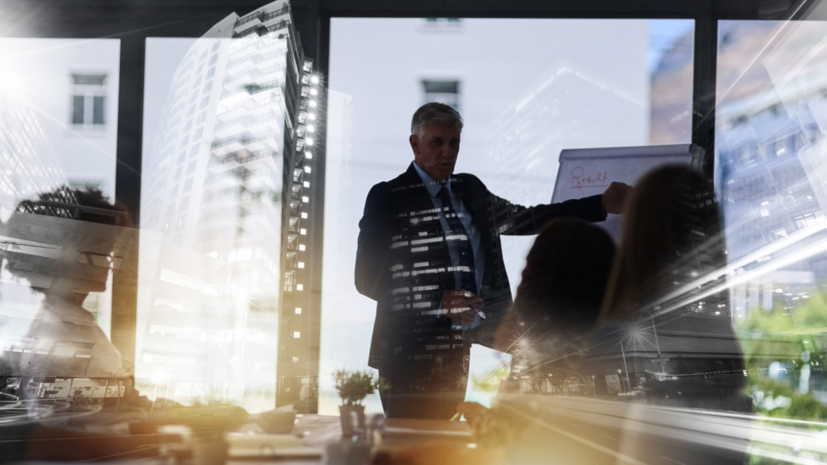 Male in office pointing at a board presenting to a group of people