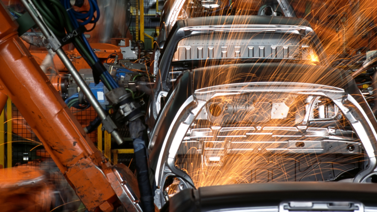 Shows cars on a factory floor being built by machines with sparks flying off them