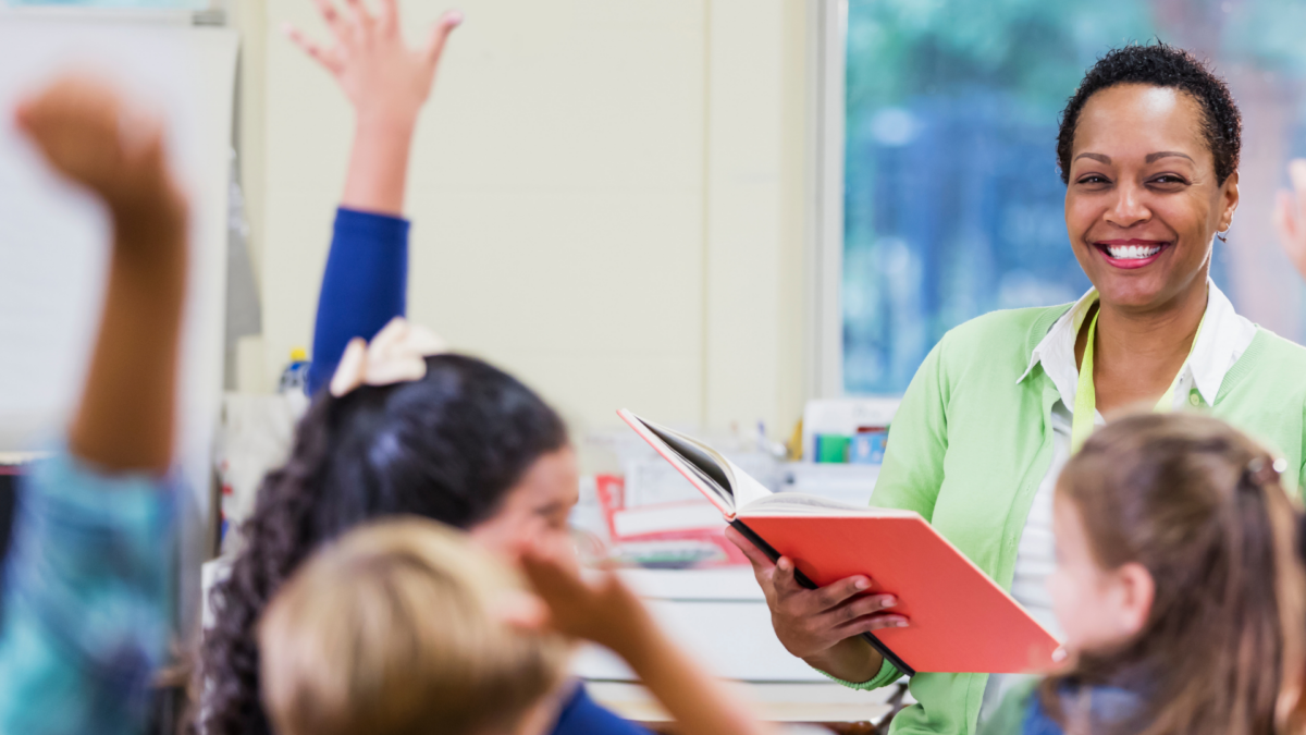 Smiling teacher at the front of the class holding a red book with pupils in front with their hands in the air