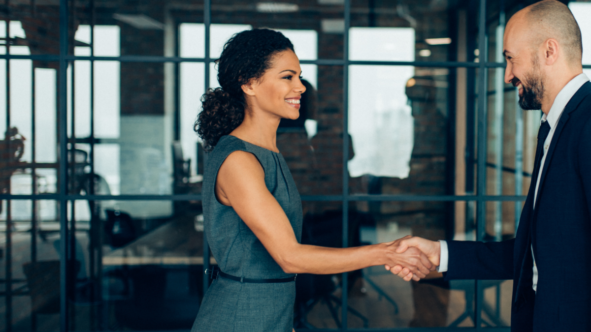 Image of a man and woman wearing business attire shaking hands and smiling, standing outside a modern styled glass fronted office building