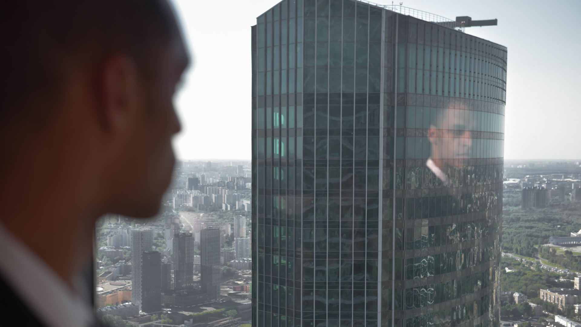 Man looking through an office window at a tall office block, his thoughtful reflection can be seen