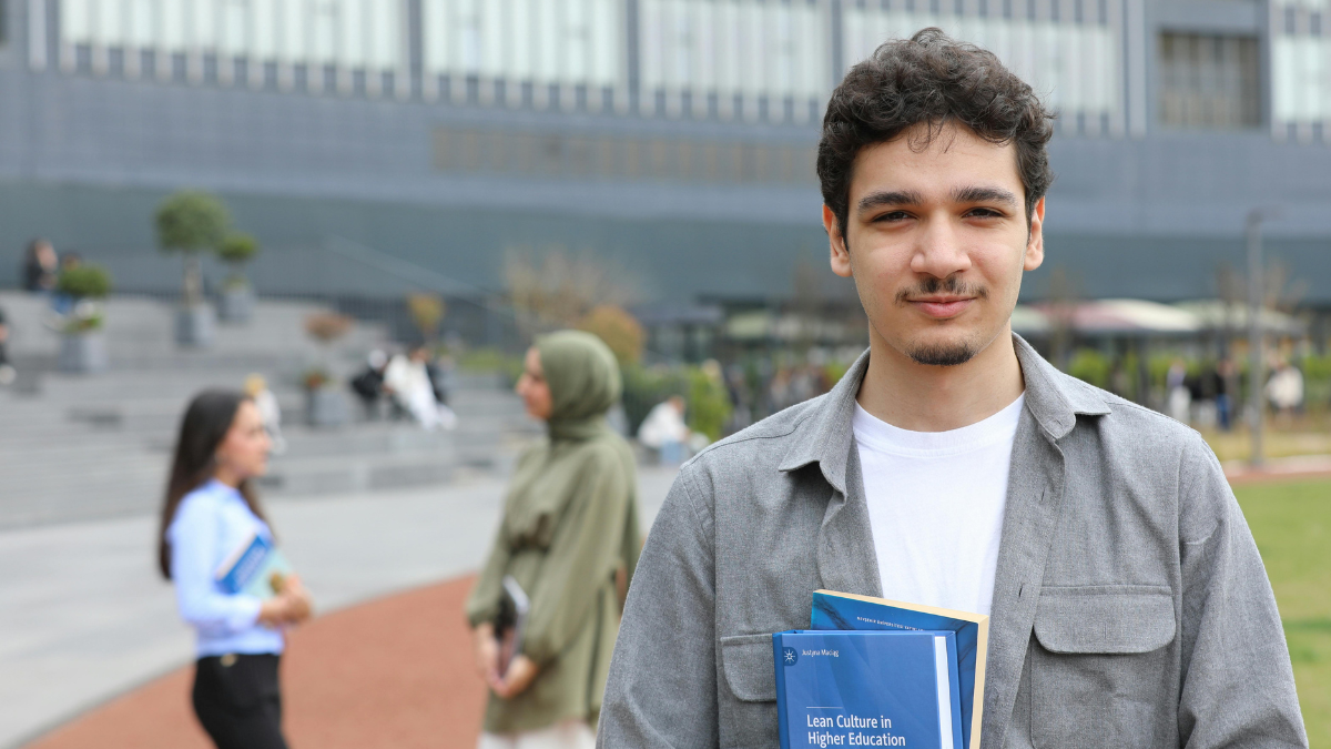 Image showing students standing outside an educational building, male in foreground holding a text book